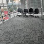 Modern office with grey patterned carpet flooring and black chairs by large glass windows on a high-rise building.